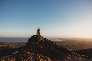 Silhouetted figure sits atop rocky mountain peak at sunset overlooking coastal landscape and ocean horizon.