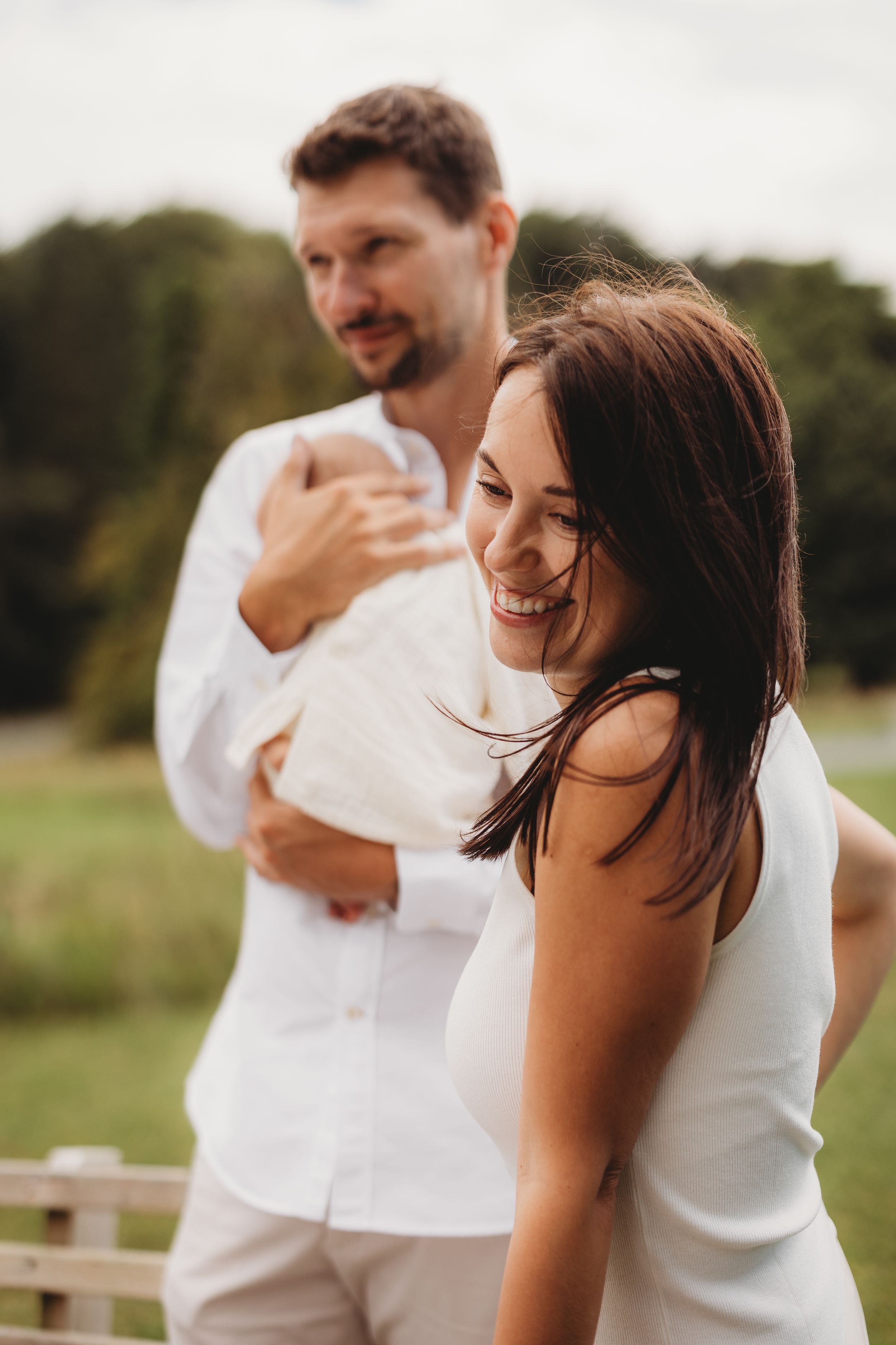 Happy couple in white outfits outdoors, man holding baby while woman smiles joyfully in a green field.