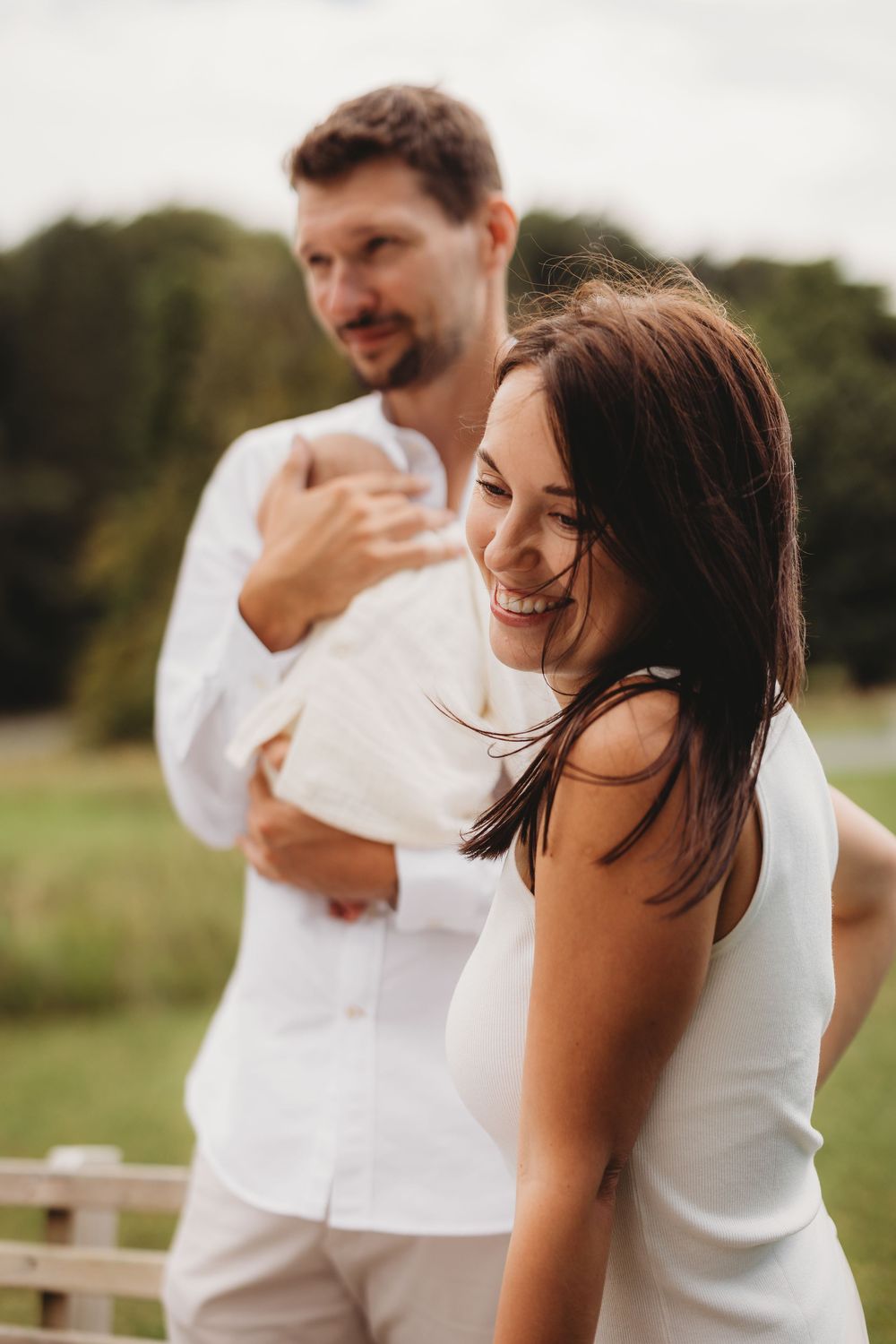 Happy couple in white outfits outdoors, man holding baby while woman smiles joyfully in a green field.