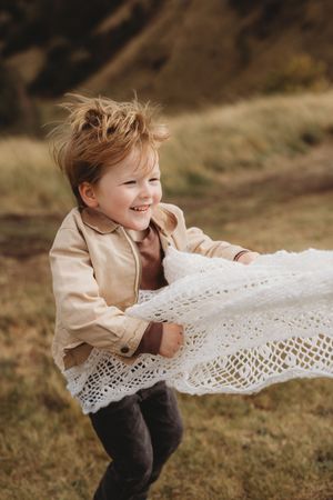 A child in a beige jacket plays joyfully with a flowing white lace fabric outdoors in a grassy field.
