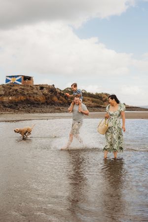 A sequence of photos showing people playing and splashing in shallow ocean water with a dog on a cloudy day at the beach.