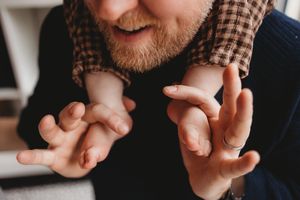 Bearded man smiling while gently holding a newborn baby's tiny hands, wearing a ring and plaid scarf.