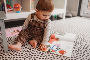 Curious baby in plaid overalls sitting on polka dot rug, exploring a colorful picture book on the floor.