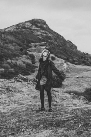 A person stands on a hillside path beneath a dramatic mountain peak in black and white photography.