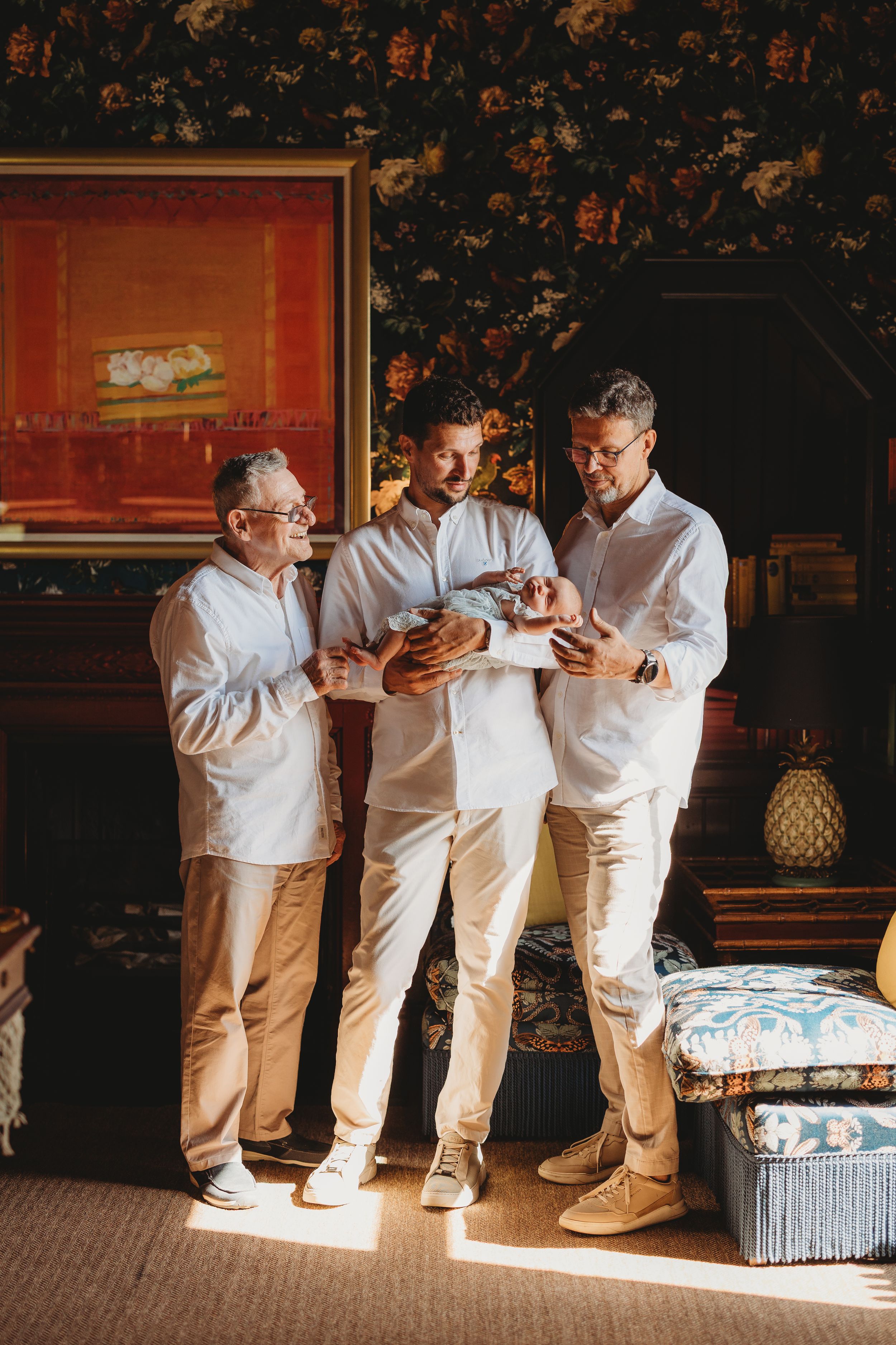 Three men in white shirts admire a newborn baby indoors, surrounded by elegant decor and warm natural light.