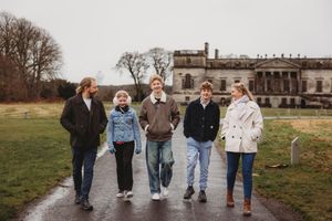 Five people walk together on a path in front of a grand historic mansion on a cloudy day.