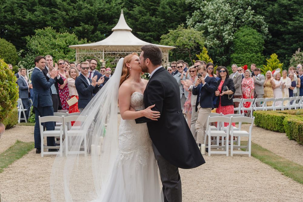 Wedding ceremony setup with white chairs and a gazebo in a lush garden setting.