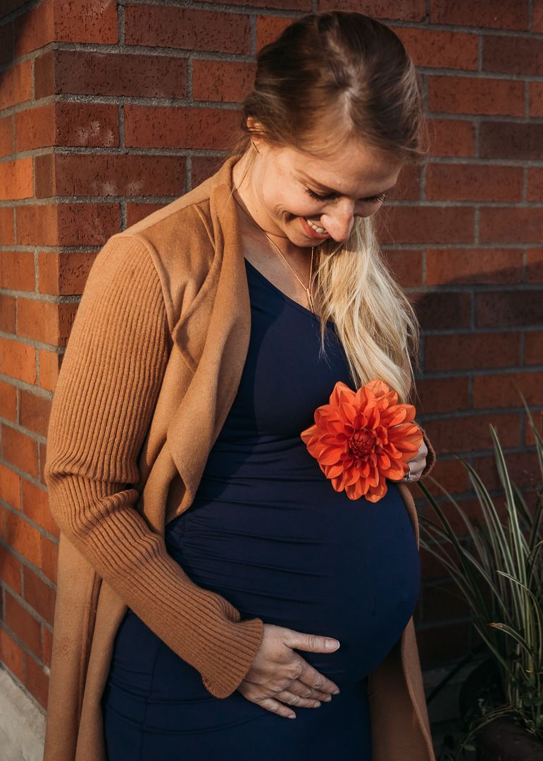 A pregnant woman in a navy dress and brown cardigan holds her belly while standing against a brick wall with an orange flower.