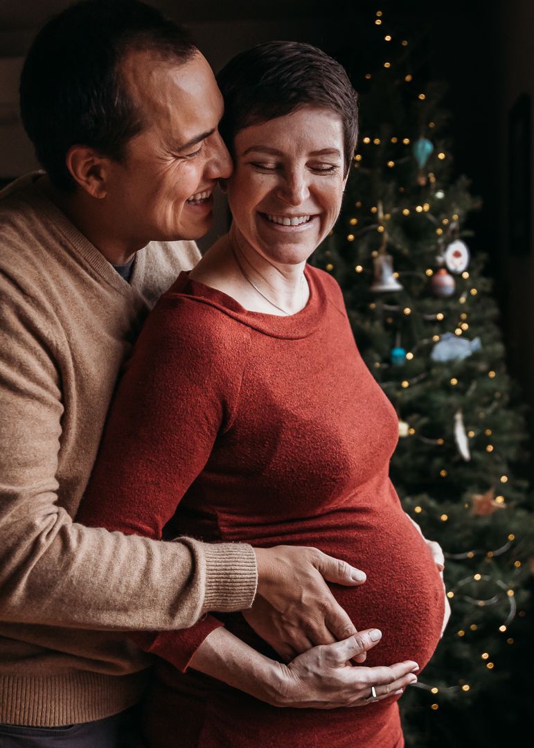 Expectant couple shares tender moments in front of a Christmas tree with twinkling lights in the background.