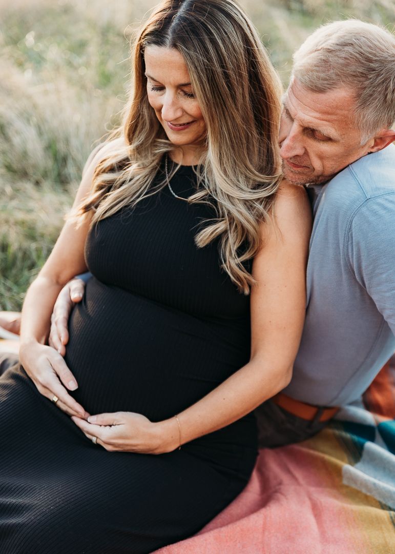 A couple shares an intimate moment outdoors while cradling a pregnant belly in a black maternity dress.