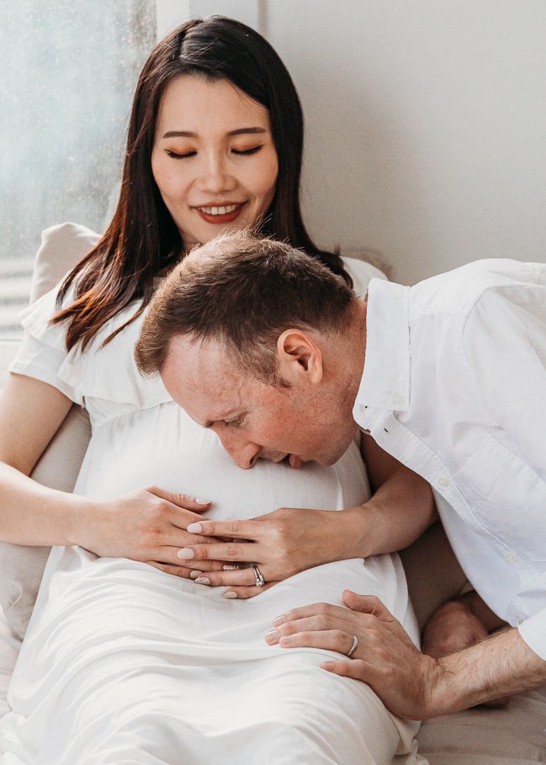 Intimate pregnancy announcement photos with couple looking at ultrasound images together on white bedding.