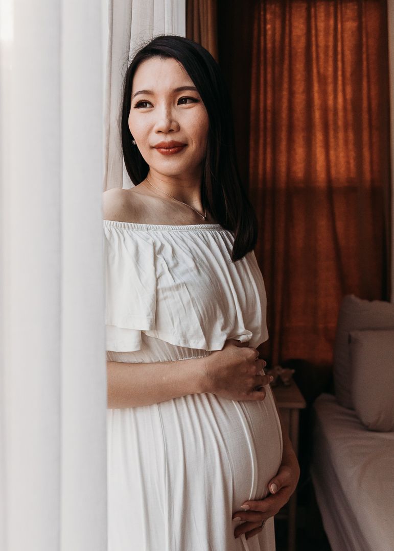 A woman in a white off-shoulder dress poses in front of brown curtains while holding her pregnant belly.