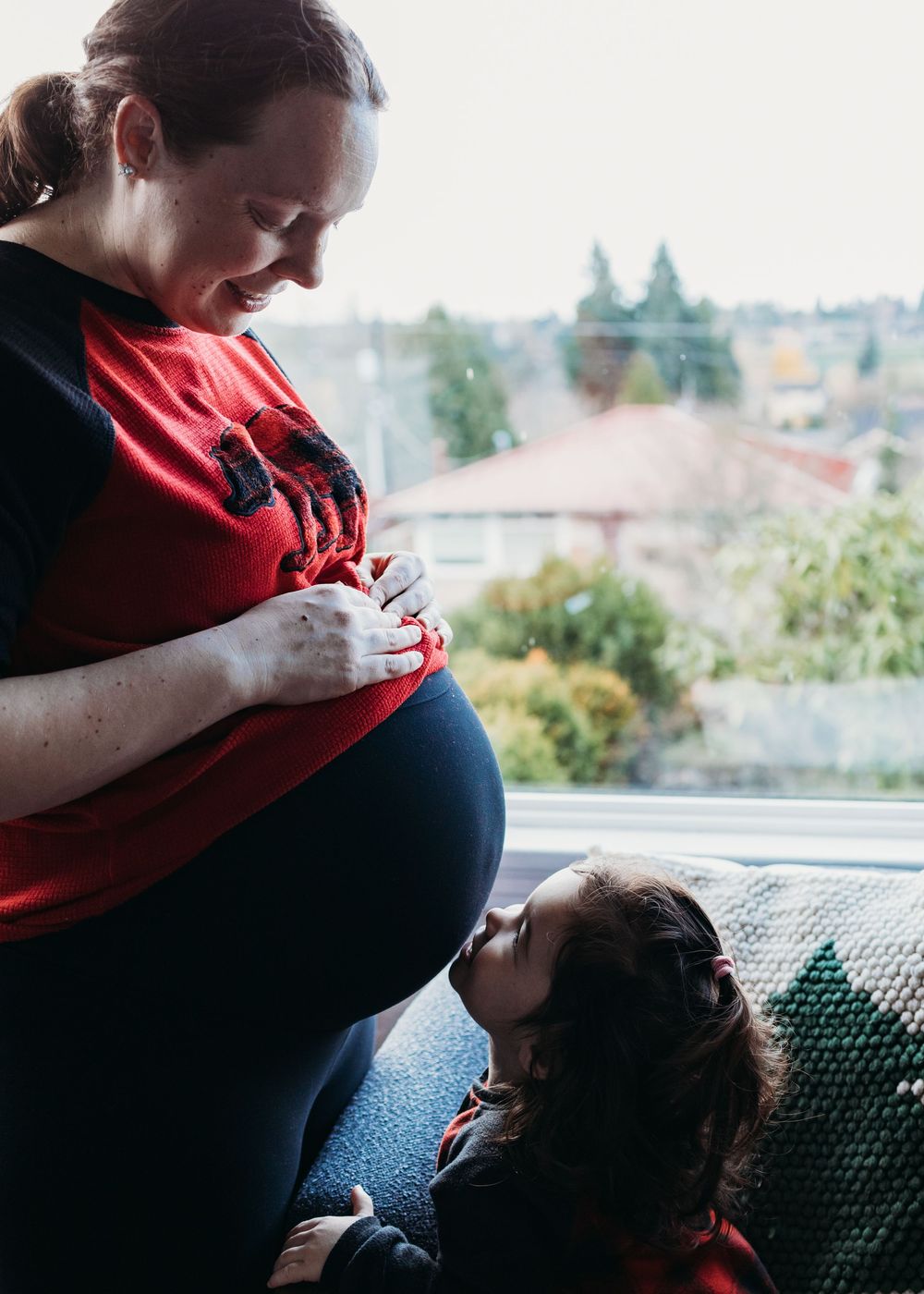A pregnant woman in a red shirt looks down at her belly while a small child gazes up at her by a window.
