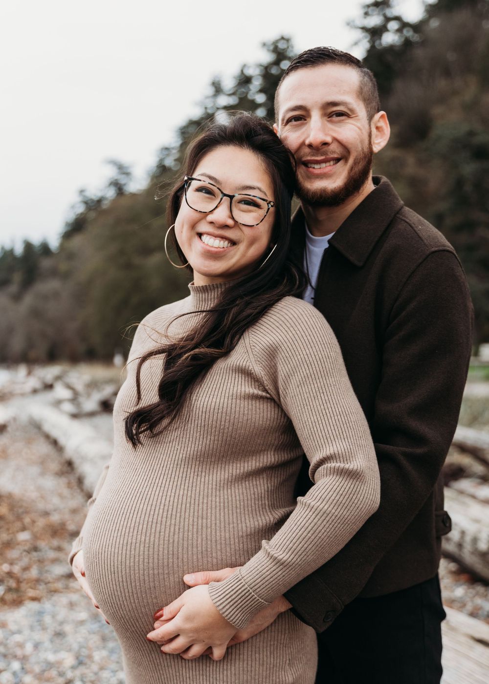 A couple embraces on a rocky beach during a maternity photoshoot, wearing cozy sweaters in neutral tones.