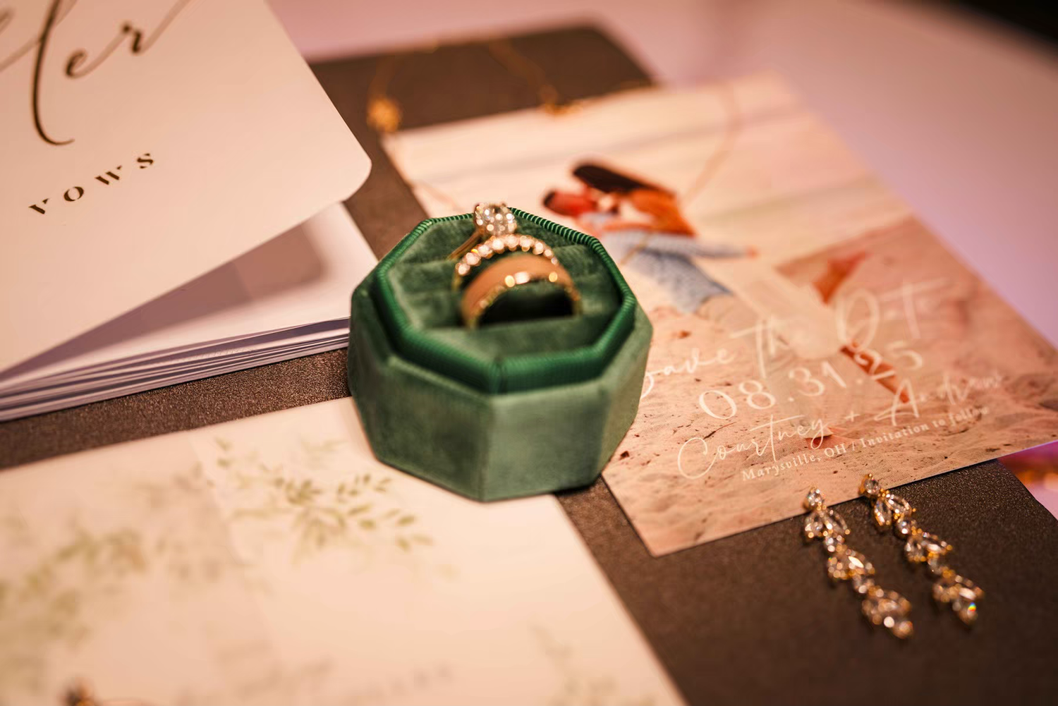 Vintage-style flatlay of wedding rings, invitations, and green velvet ring boxes arranged on a pink surface at Retreat 21 Wedding Venue