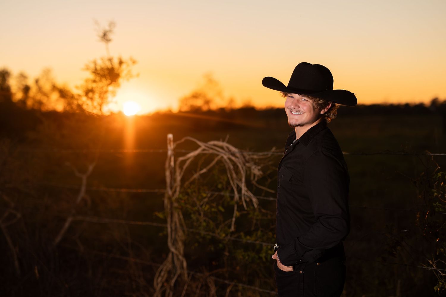 Silhouette of person in cowboy hat against dramatic sunset in rural field.