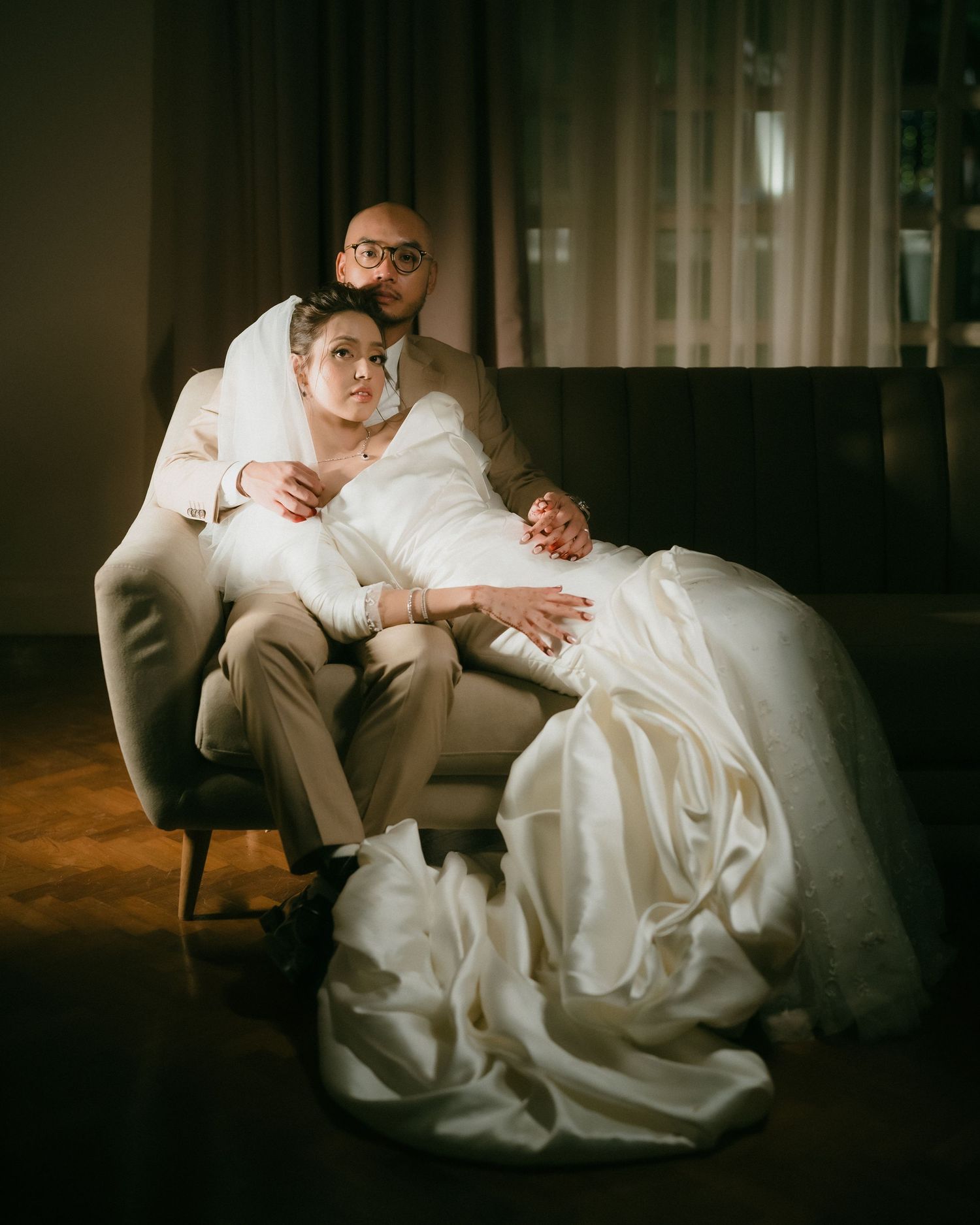 Bride and groom posing intimately in a dimly lit room, bride in a white gown sitting on a chair.