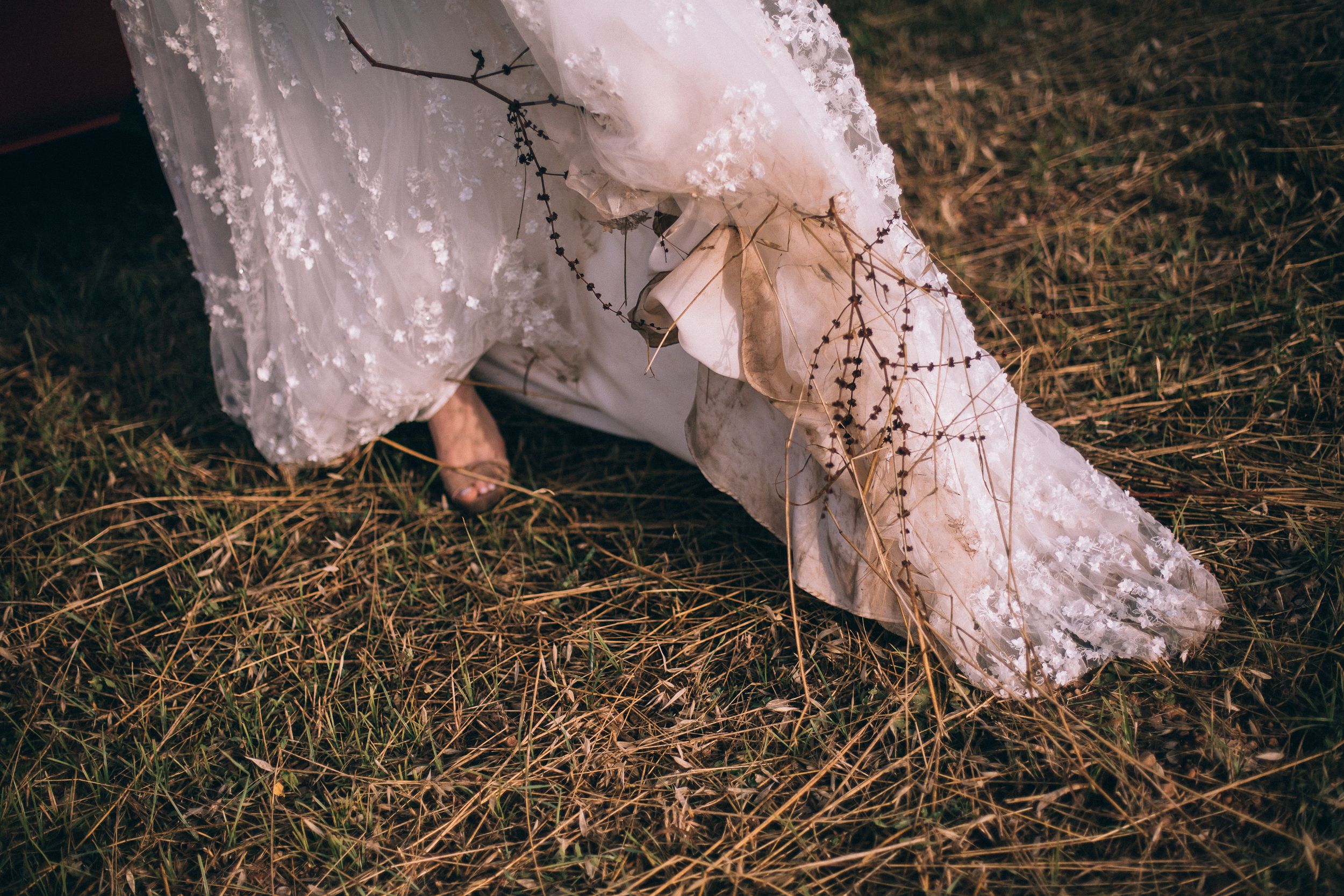 Close-up details of a delicate lace wedding dress train trailing across rustic grass and ground.
