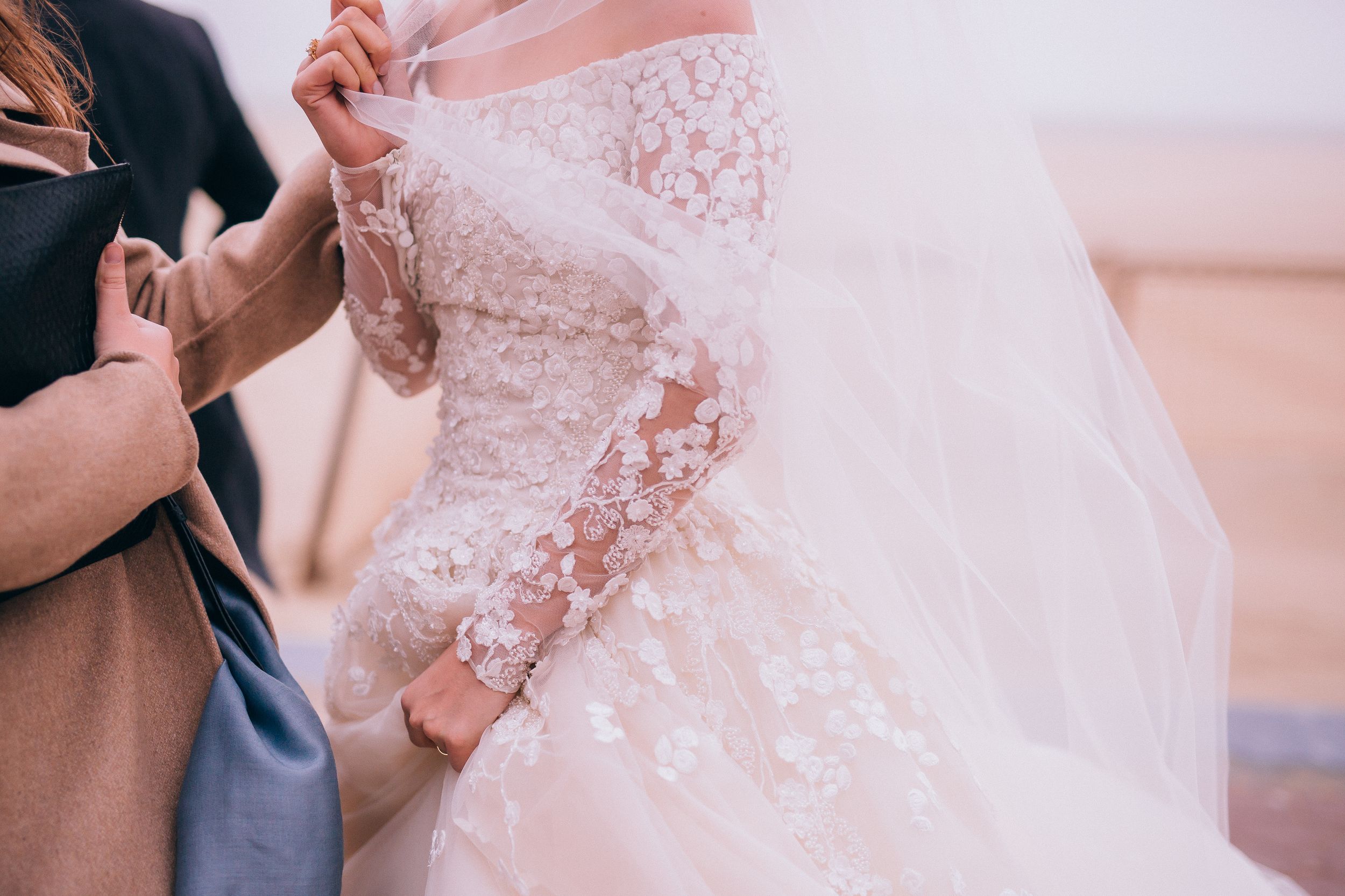 Bride in elegant lace long-sleeve wedding gown with floral appliques being assisted by a helper outdoors.