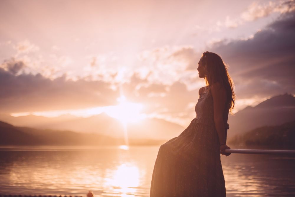 Silhouetted figure stands on beach at sunset with dramatic orange sky and mountains in background.