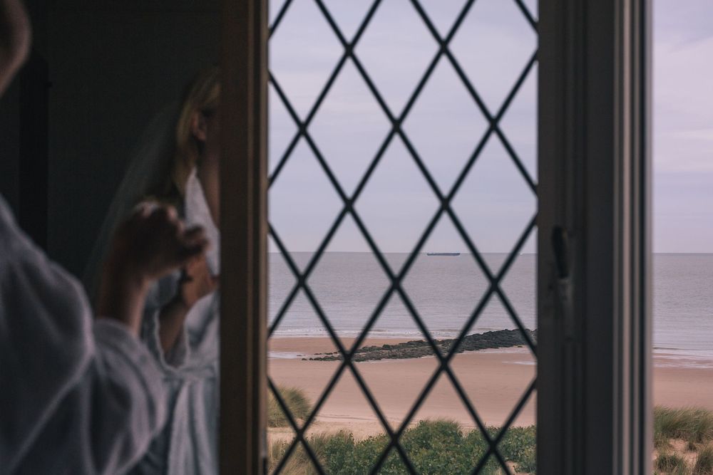 Person sitting by a window with diamond-patterned glass panes overlooking a coastal landscape at sunset.