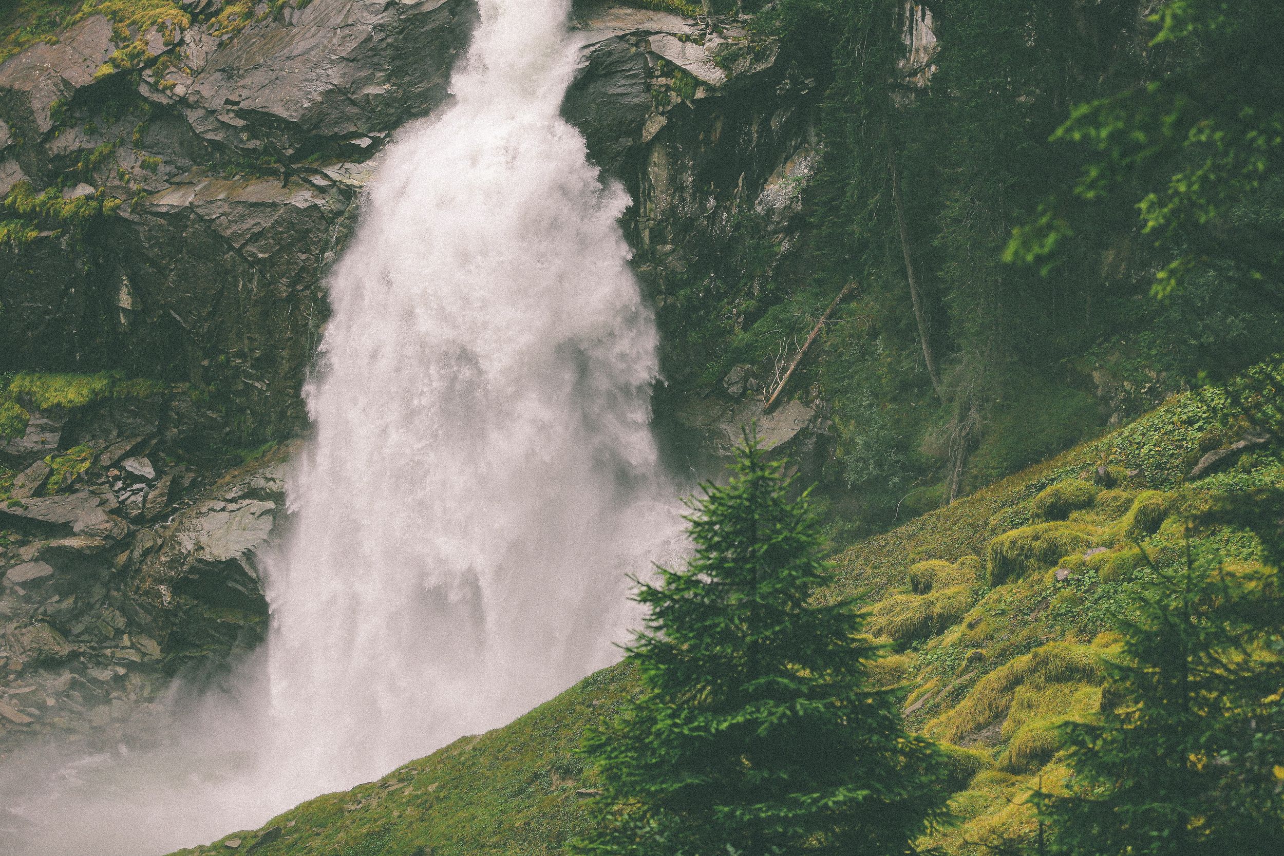 A powerful waterfall cascades down a rocky mountain cliff surrounded by lush green trees and moss-covered slopes.