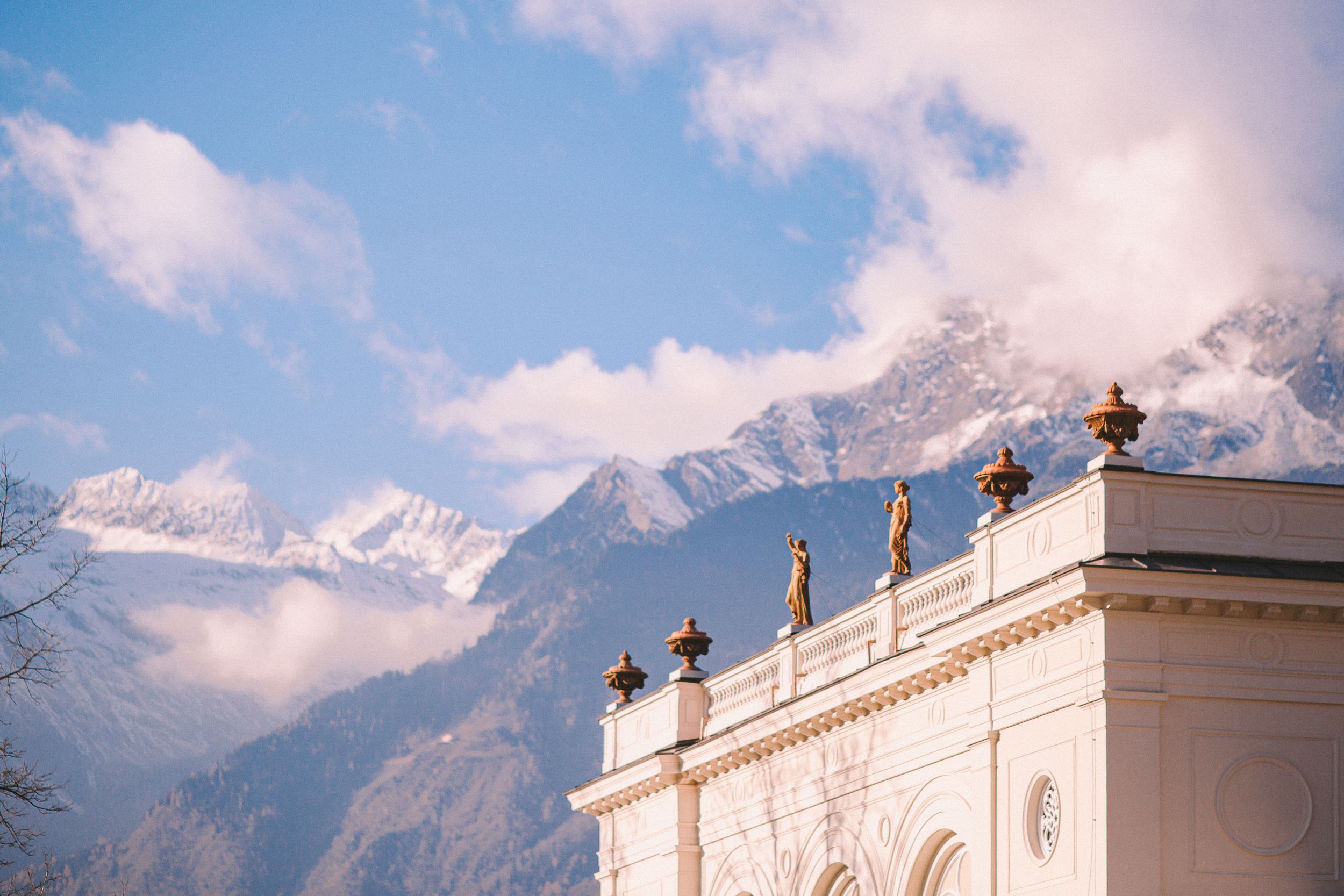 White neoclassical building with golden statues on rooftop against snow-capped Alpine mountains and blue sky.