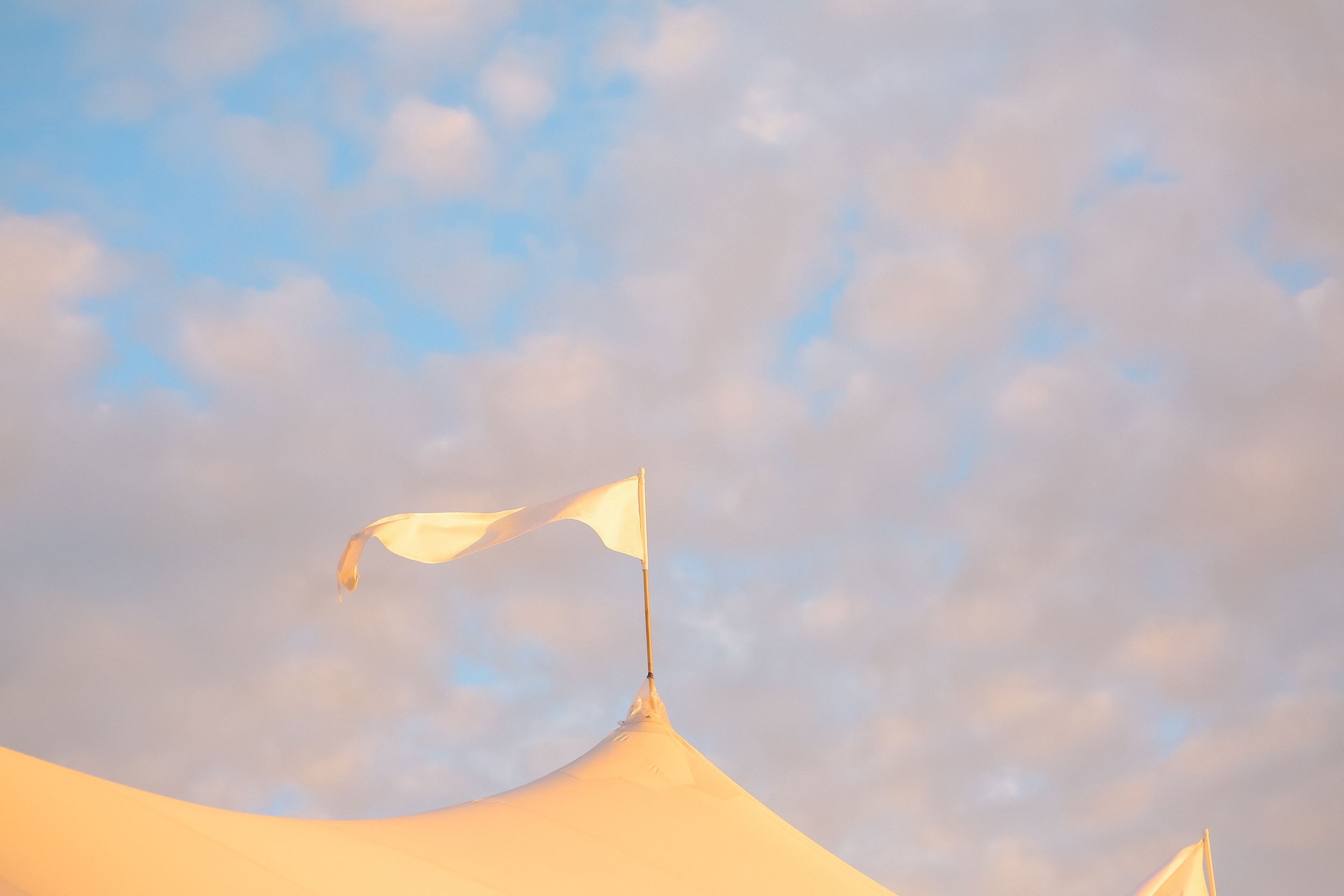 White circus tent top with a flag against a soft blue cloudy sky at golden hour.