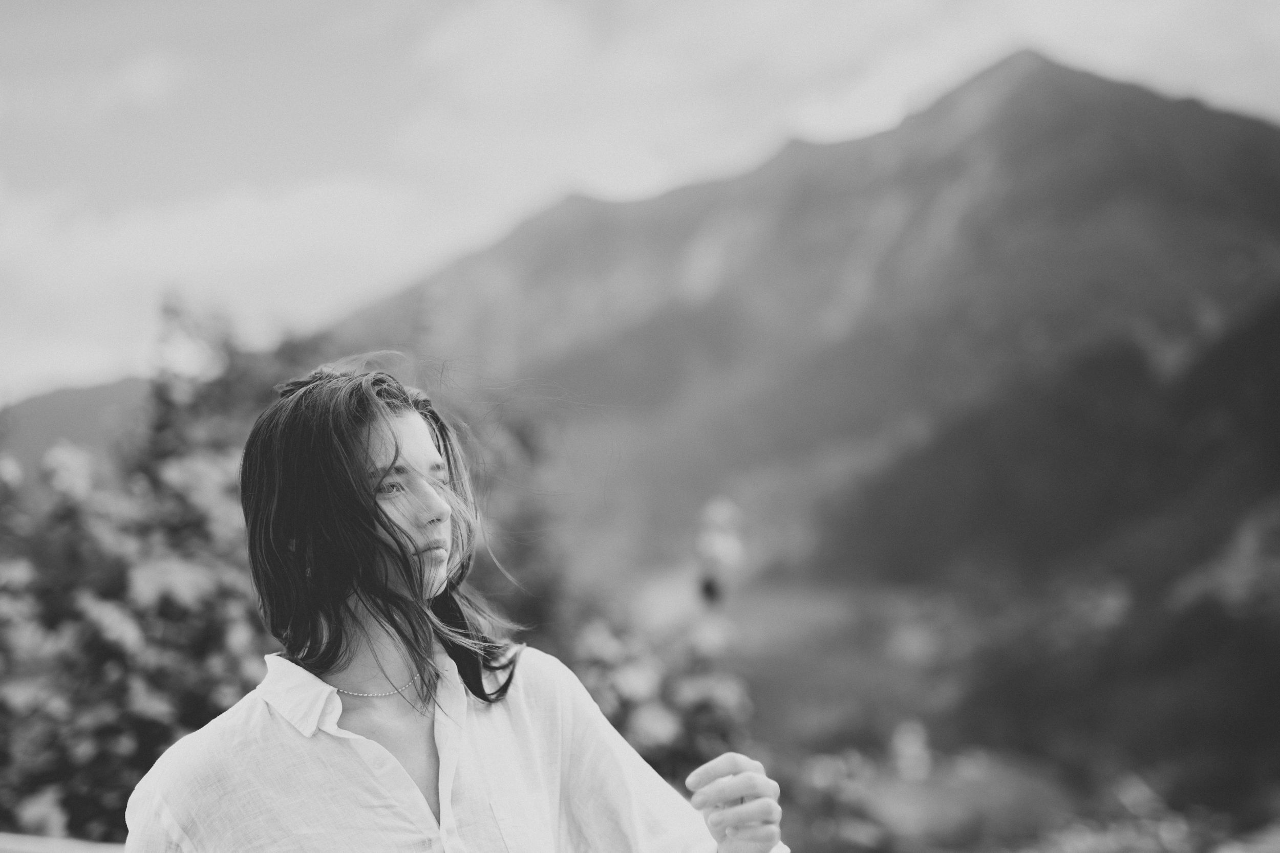 Black and white photo of a person in white clothing standing on a mountain overlook with wind-blown hair.