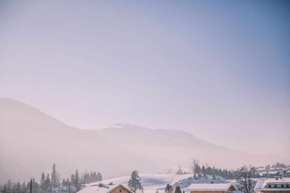 Scenic mountain landscape with evergreen trees and snow-capped peaks against a misty blue sky at sunset.