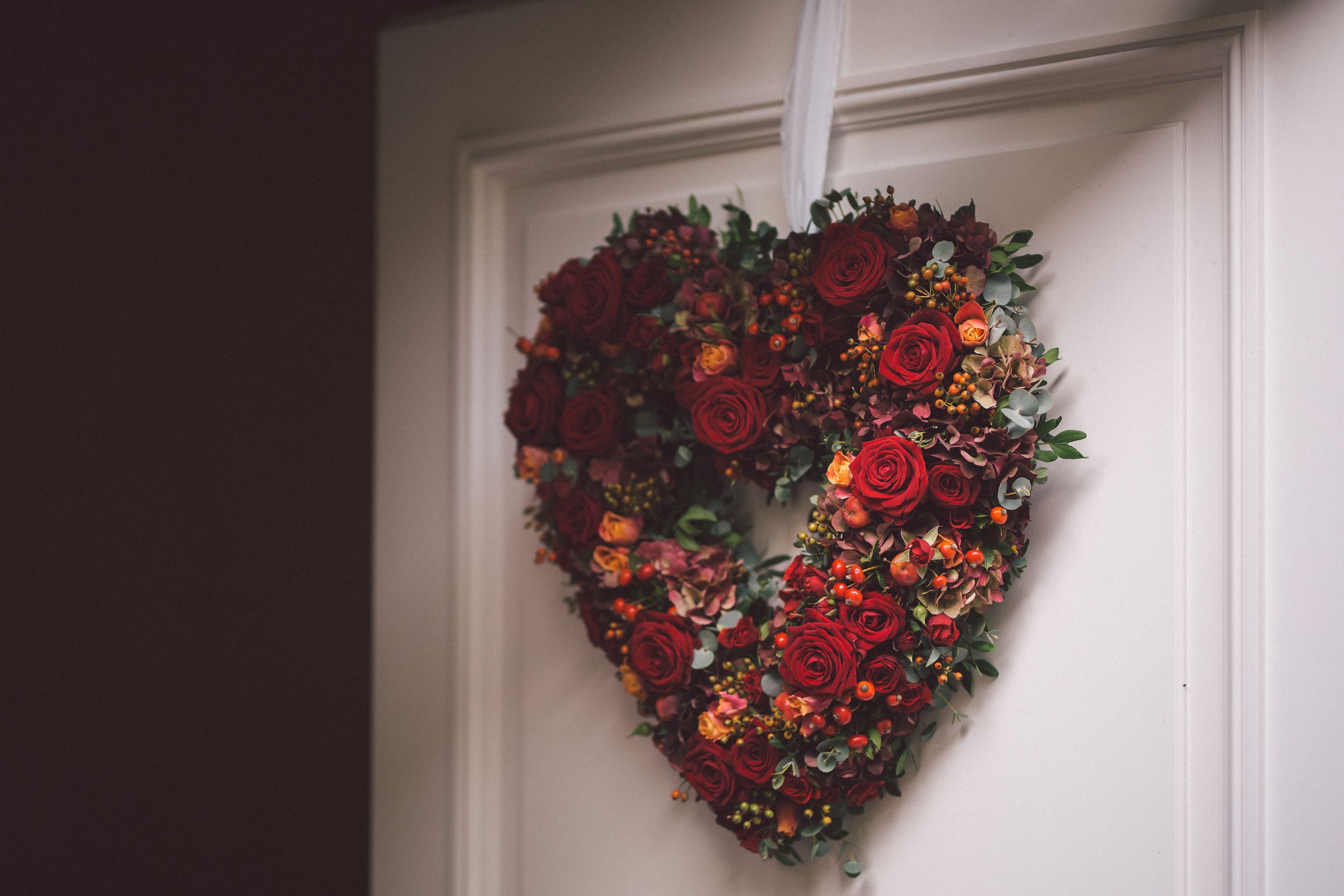 A heart-shaped red and orange floral wreath with berries and autumn foliage mounted on a white door.