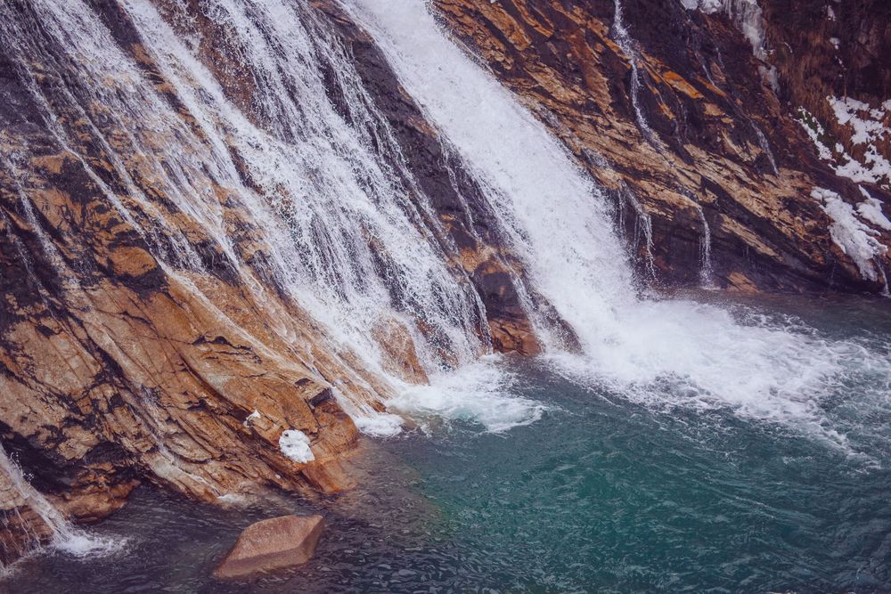 Scenic waterfall cascades down rocky cliffs into turquoise pool surrounded by rugged mountain terrain.