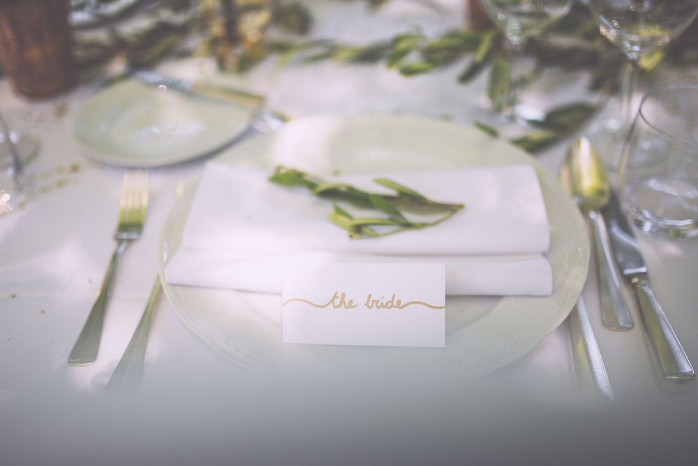Wedding place cards with greenery decorations and calligraphy sit elegantly on a white tablecloth.
