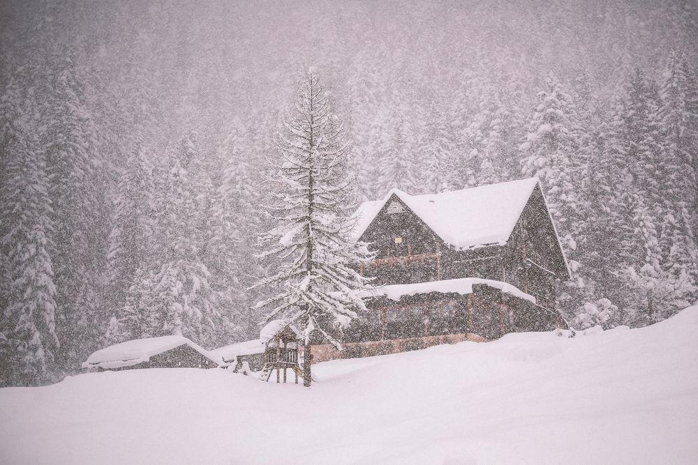Snow falls heavily on rustic wooden cabins and evergreen trees in a serene winter mountain landscape.