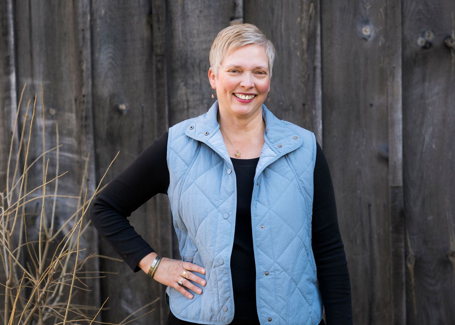Person wearing a light blue quilted vest and black long sleeve shirt poses with hand on hip against wooden fence.