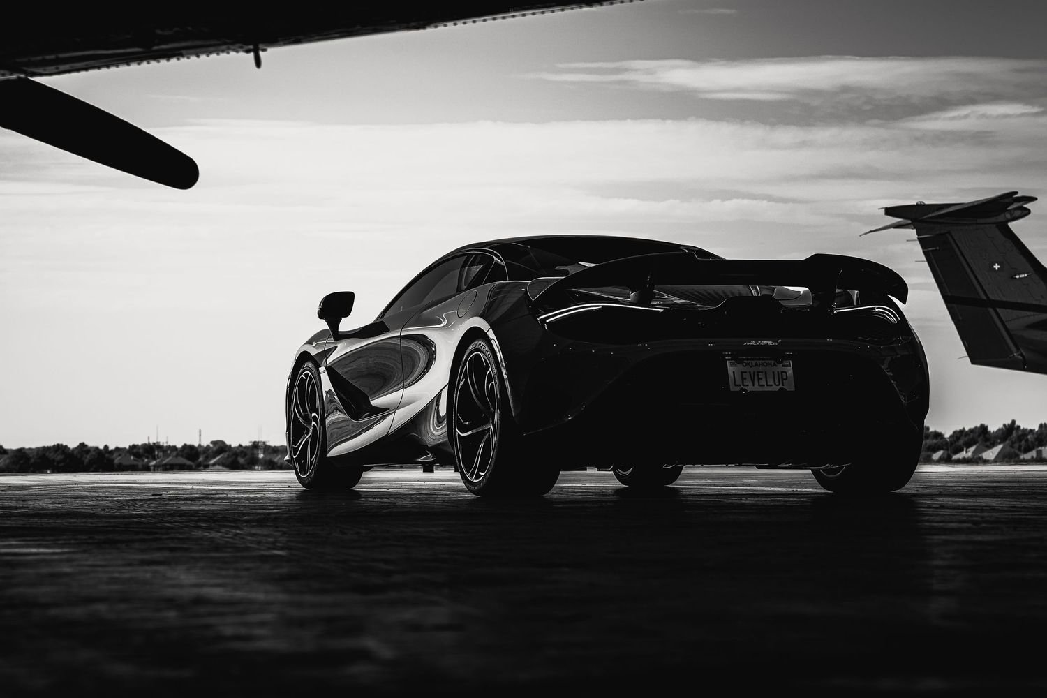 A black McLaren supercar photographed from behind at sunset on an airfield in dramatic black and white.