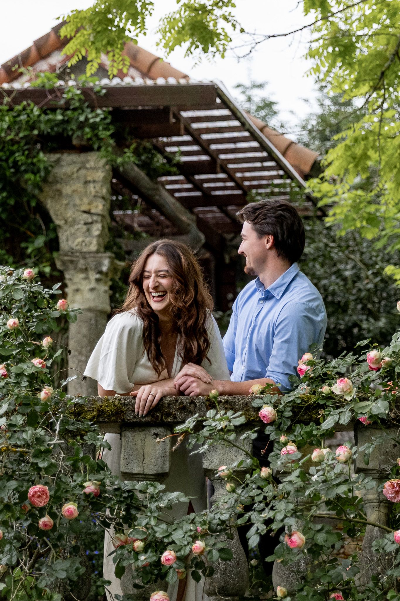 Couple laughing together leaning on a stone wall covered in pink climbing roses.