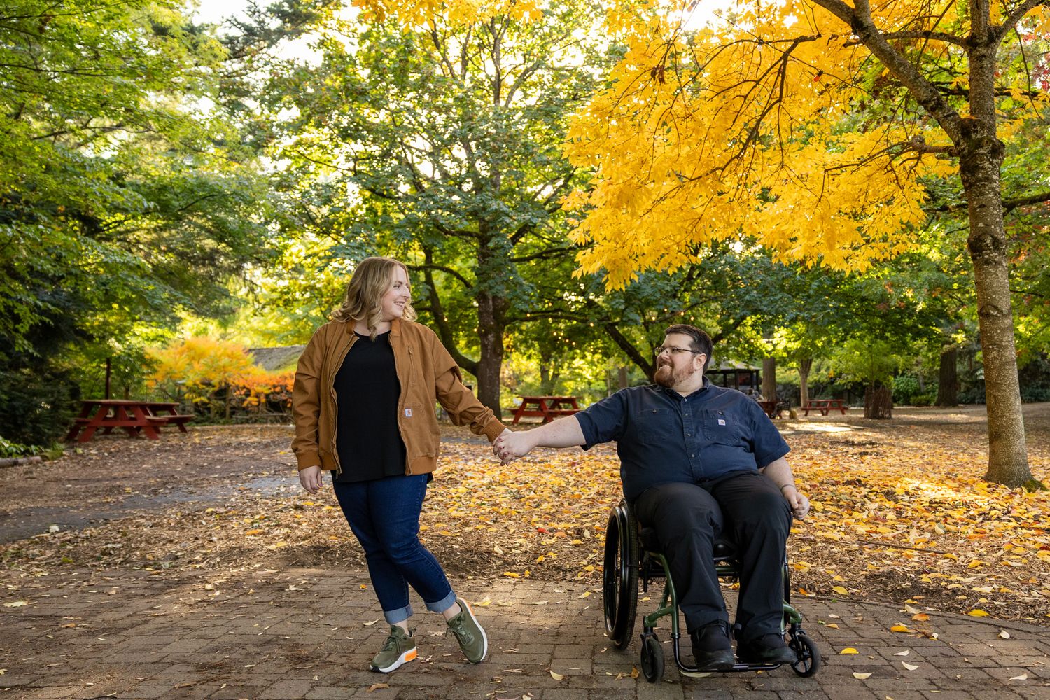 Couple holding hands in autumn park, woman walking beside man in wheelchair among golden trees.
