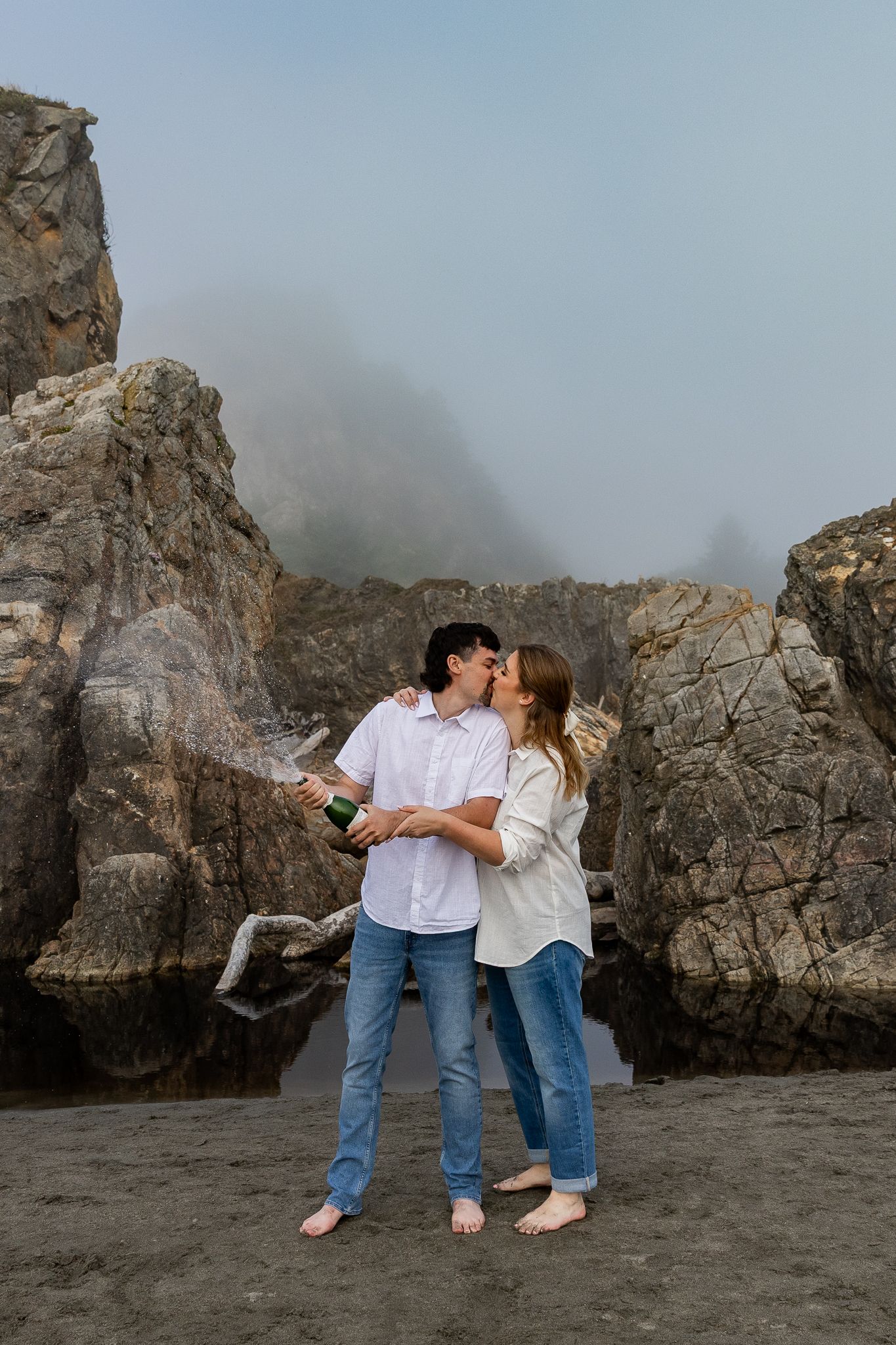 Couple kissing while popping champagne bottle on a rocky foggy beach during engagement shoot.