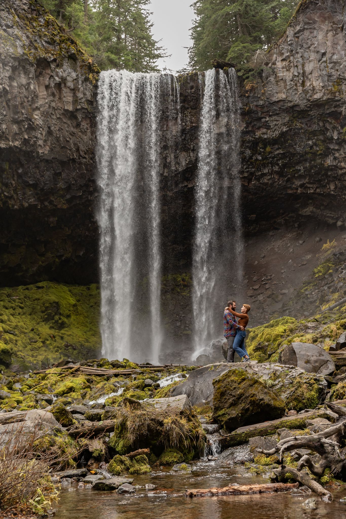 Couple embracing in front of a tall waterfall surrounded by mossy rocks.