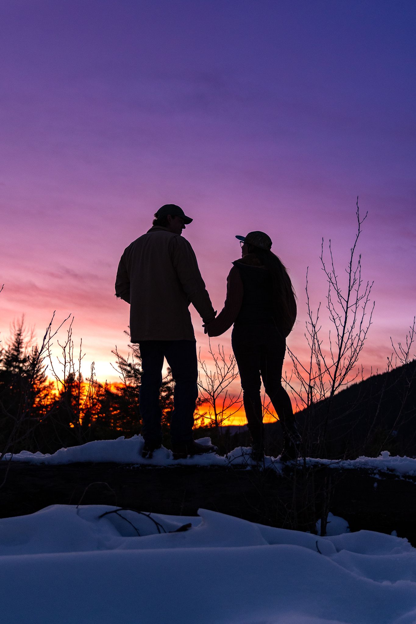 Romantic silhouette of couple holding hands on snow-covered log during stunning colorful winter sunset.