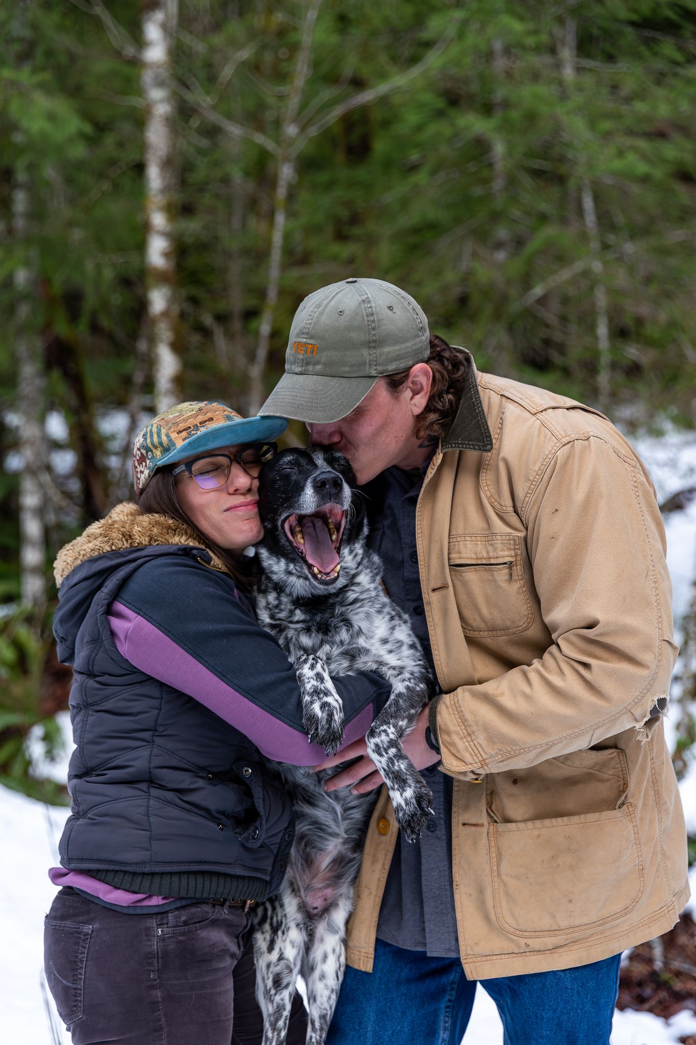 Couple kissing their happy spotted dog in a snowy winter forest setting.