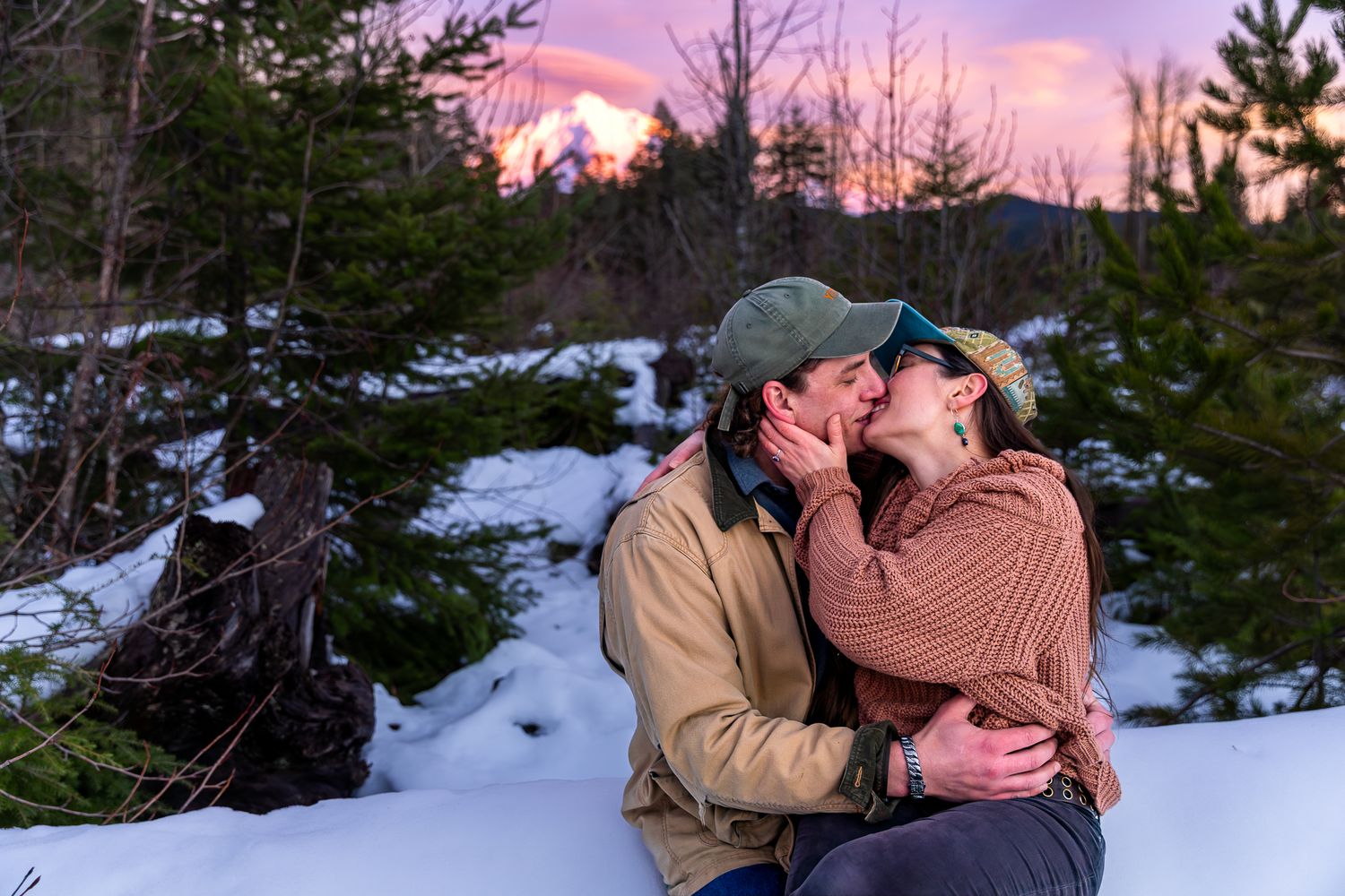 Couple sharing a romantic kiss in a snowy forest with a vivid pink and purple sunset sky.