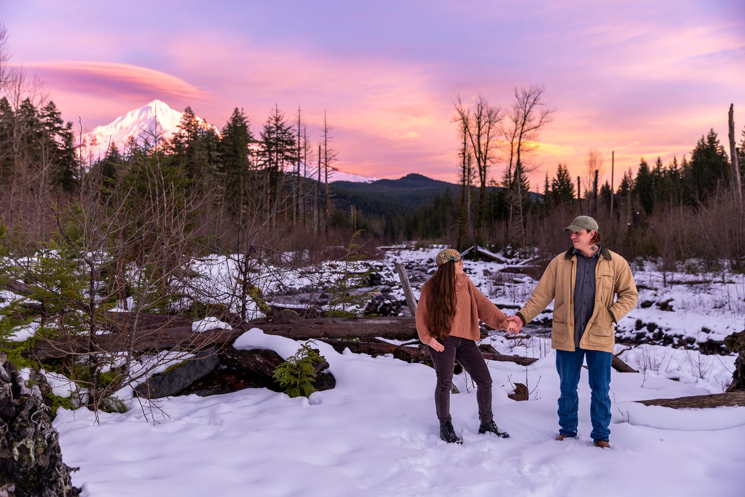 Couple standing in a snowy landscape with Mount Hood and a stunning pink sunset sky behind them.