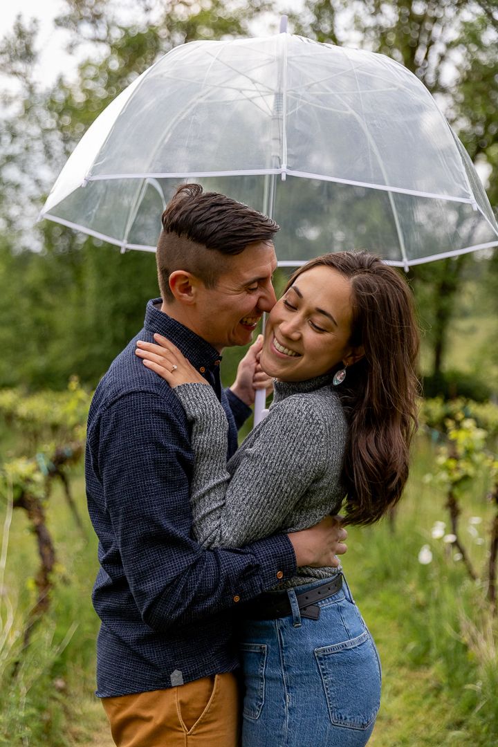 Couple sharing a tender romantic moment embracing under a clear bubble umbrella in green vineyard.