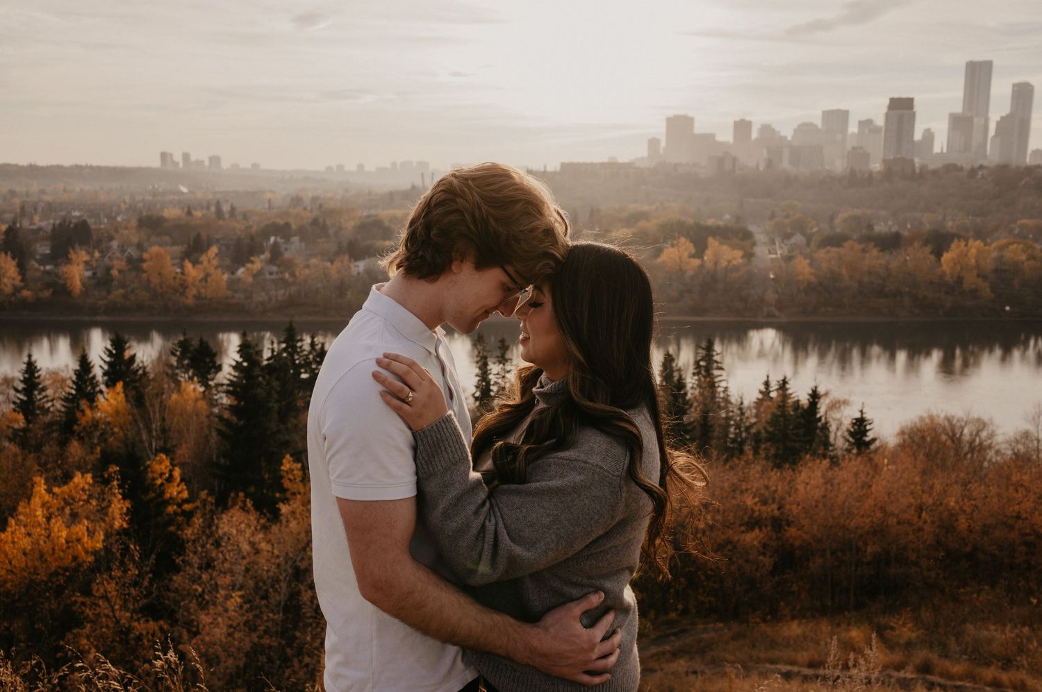 Couple embraces on a hilltop overlooking a scenic autumn landscape with city skyline in the distance.