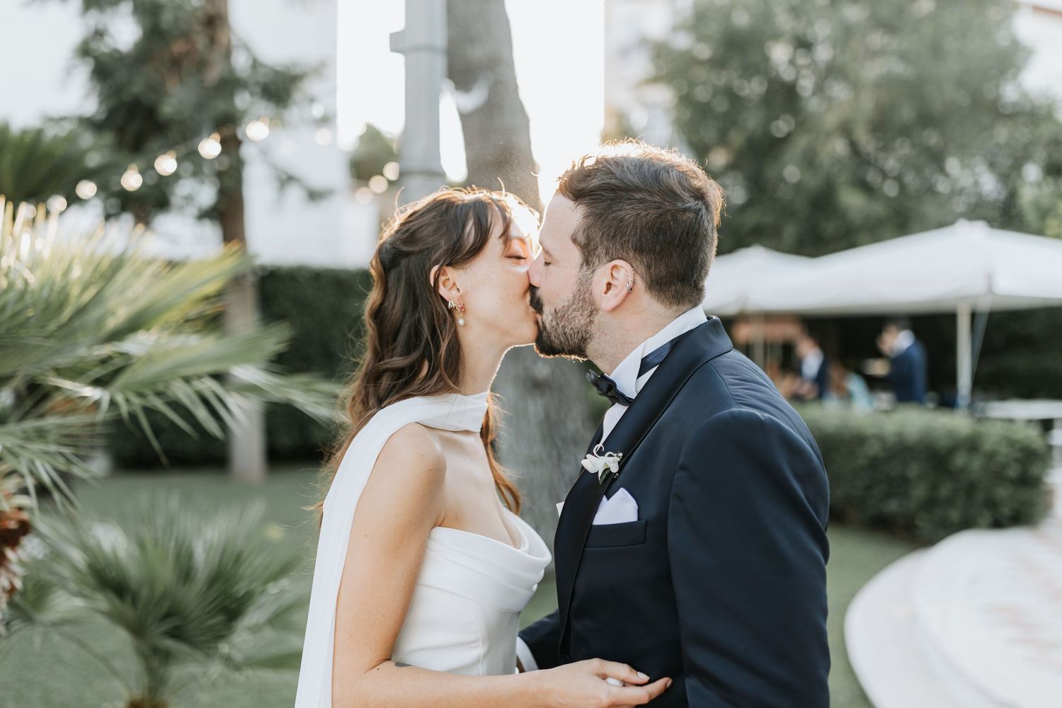 A romantic wedding couple shares a tender kiss in a garden setting with palm trees and white tents in the background.