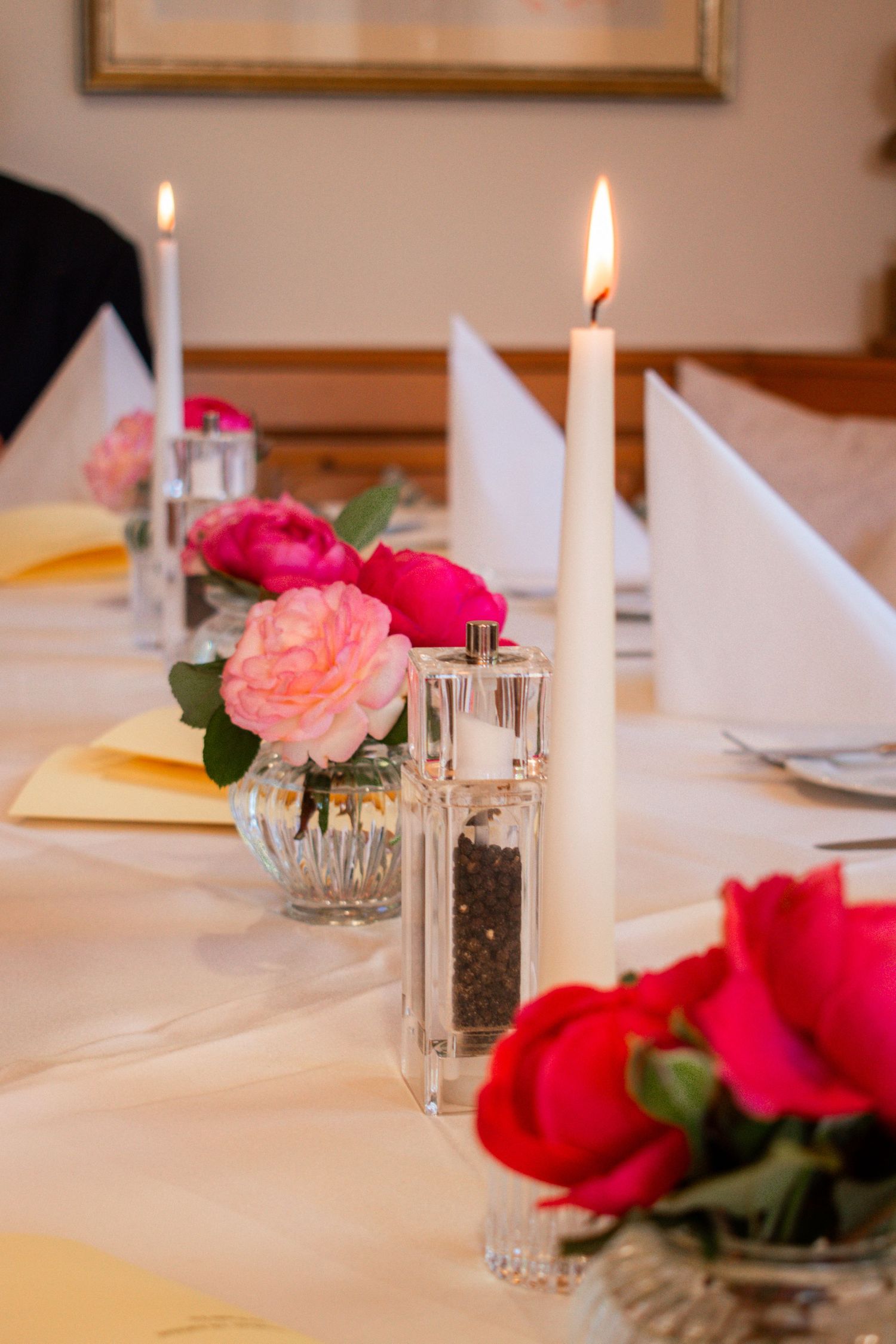 Elegant table setting with lit candles and pink and red roses in glass vases on white linens.