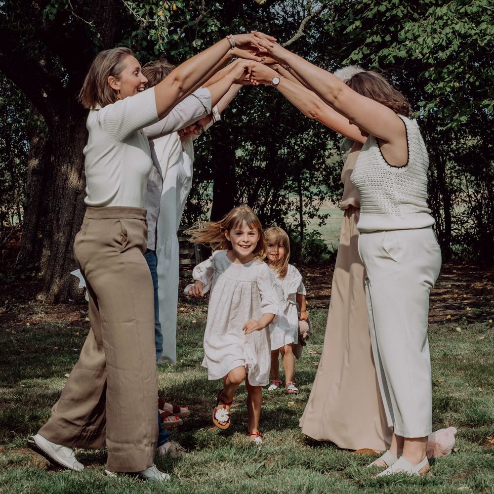 Two women forming an arch with their arms while two young girls run underneath in a sunny park.