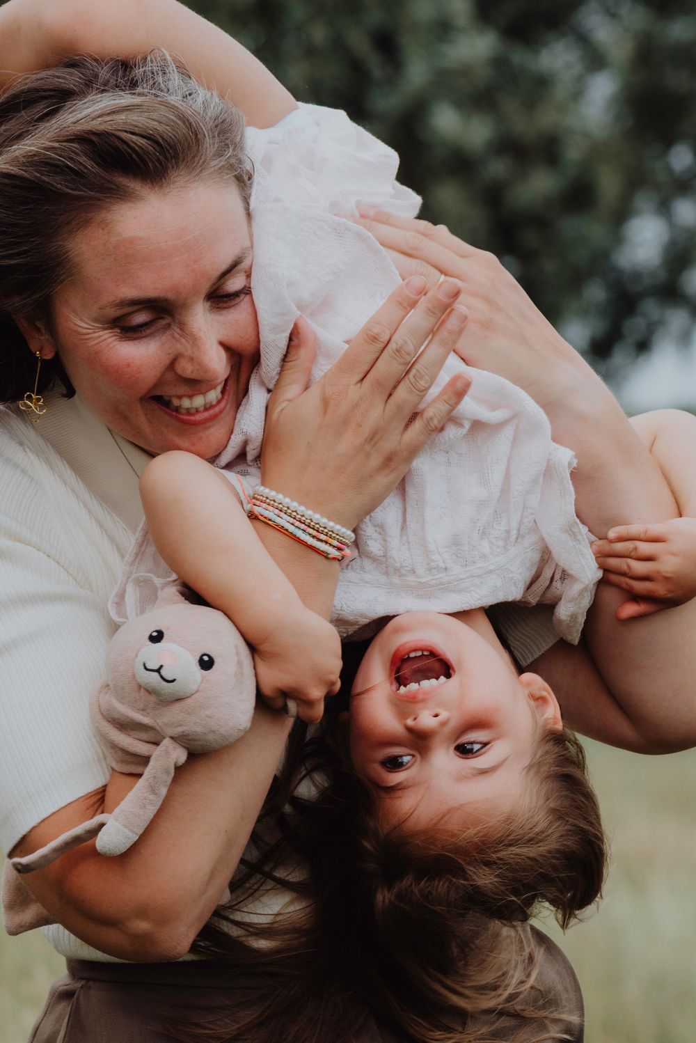 Smiling mother playfully holding her laughing toddler upside down while clutching a stuffed bunny toy.